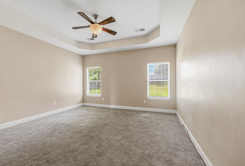 Spacious room featuring a tray ceiling, ceiling fan with chandelier lighting, two windows with horizontal blinds, neutral-toned walls, and gray carpet flooring