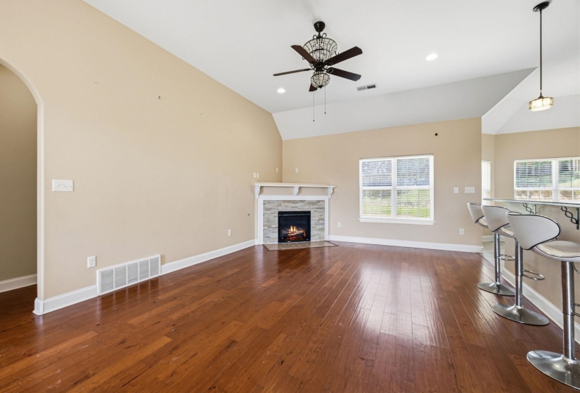 Spacious living area featuring wood-finish flooring, a stone-surround fireplace with mantel, and a ceiling fan with decorative light fixture