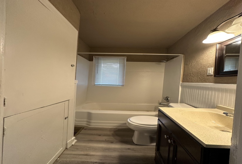 Bathroom featuring a white bathtub and shower surround, wood-finish flooring, a toilet, a dark wood-finish vanity with an integrated sink, and wainscoting