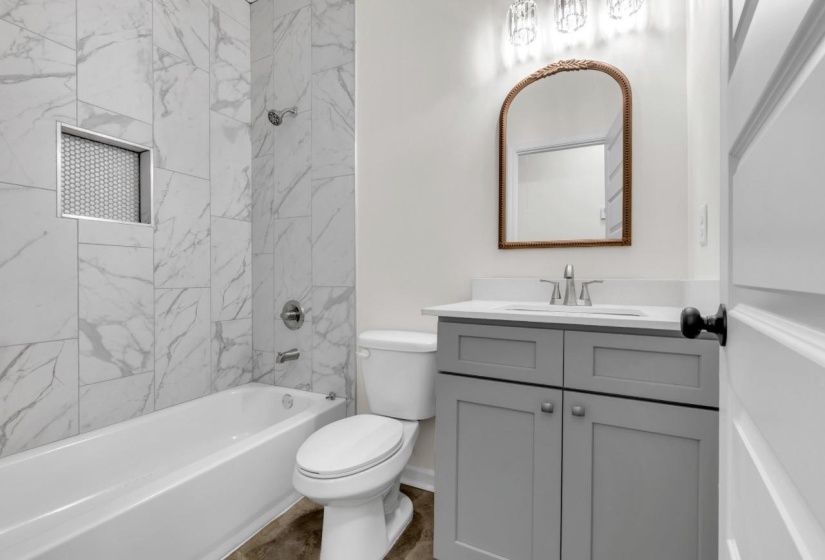 Full bathroom featuring a white porcelain tub, large-format marble-look tile surround with a recessed niche, a gray vanity with a white countertop and undermount sink, and an arched mirror with a decorative frame