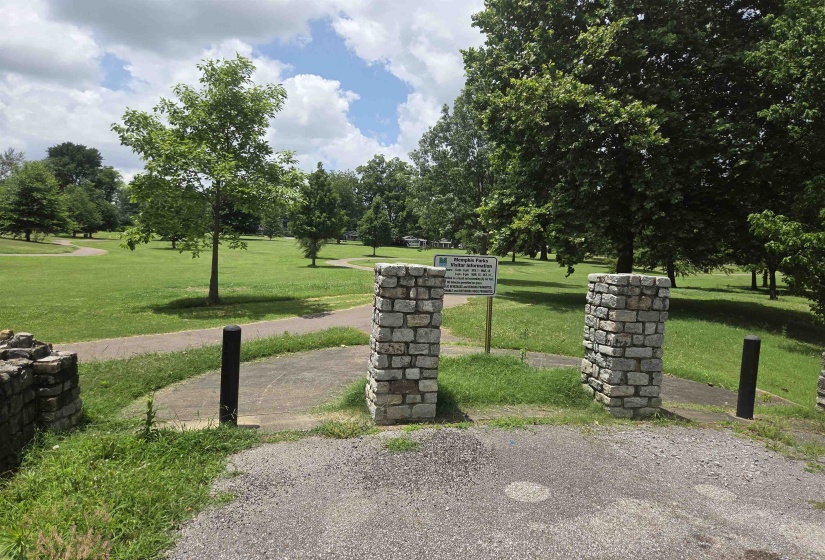 Stone column entrance with a gravel and paved pathway leading to an expansive green space with mature trees and a walking path