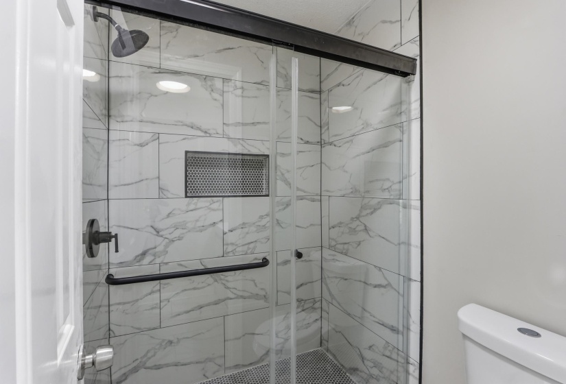 Glass-enclosed shower featuring large format white tile with grey veining, matte black shower fixtures, a built-in shower niche with a perforated metal insert, and a black-framed sliding shower door
