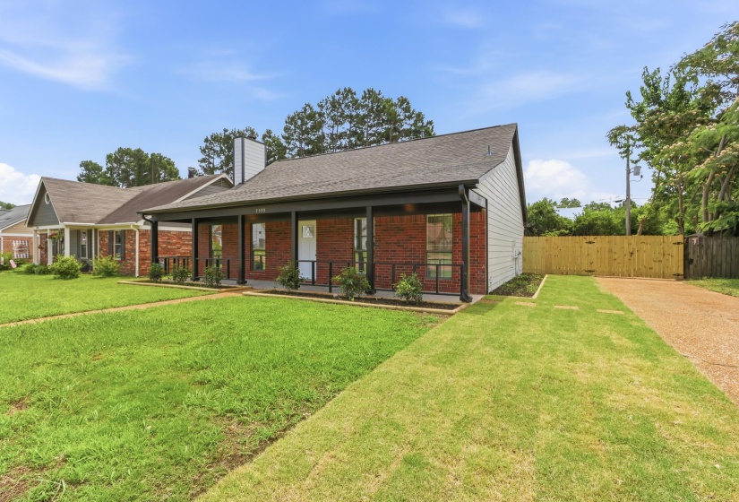 Brick facade residence featuring a covered front porch with dark columns and railing, white siding, a gravel driveway, and a wooden privacy fence