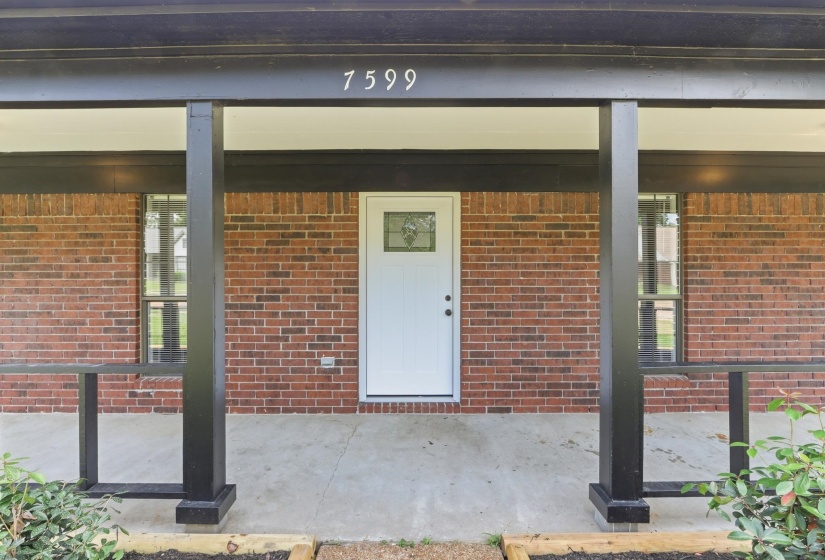 Covered front porch featuring brick siding, a white entry door with decorative glass, dark support columns, and concrete flooring