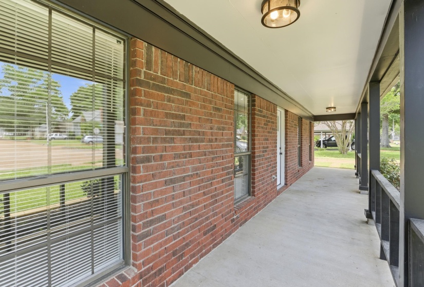 Expansive covered porch featuring a concrete surface, brick exterior, multiple windows with blinds, a white entry door, and recessed lighting fixtures