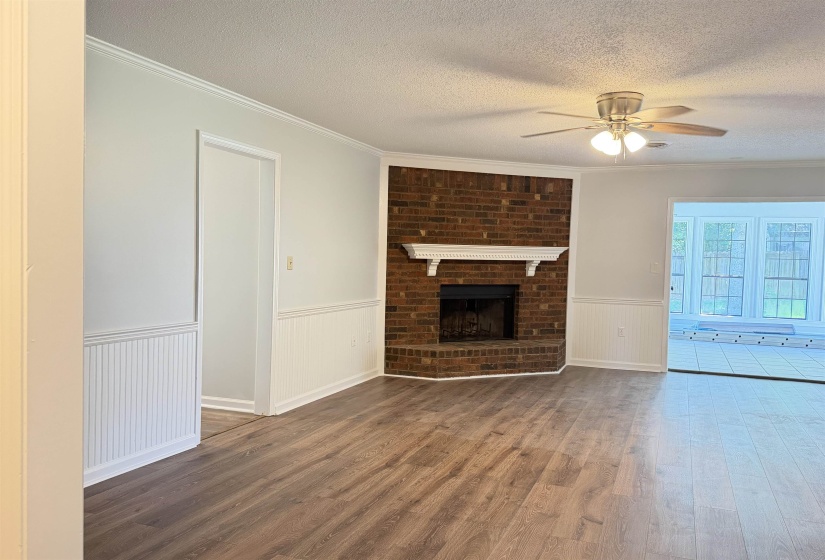Living area featuring wood-finish flooring, a brick fireplace with a white mantel, white beadboard wainscoting, crown molding, and a ceiling fan with light fixture