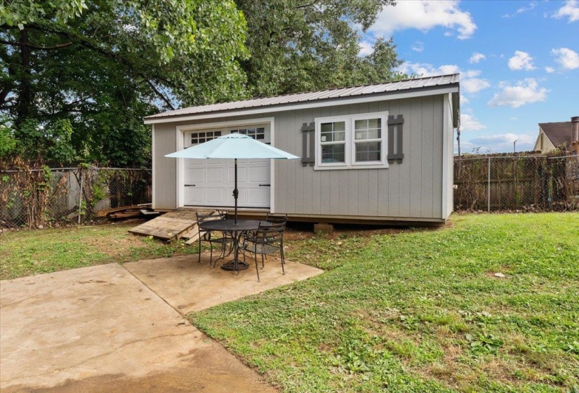 Detached shed structure featuring a metal roof, two windows with decorative shutters, and a roll-up garage door