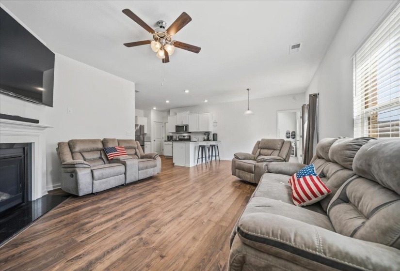 Open-concept living area featuring wood-finish flooring, a ceiling fan with integrated lighting, and a white mantel fireplace with a dark hearth