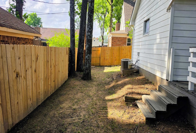Private backyard featuring a wood privacy fence, existing mature trees, a white-sided exterior, a ground-level air conditioning unit, and exterior wooden steps