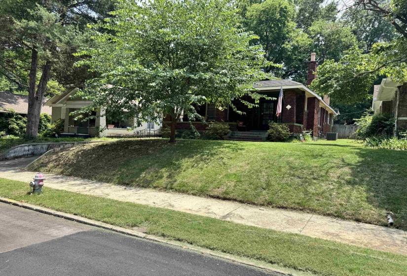 Brick facade residence featuring a covered front porch, prominent chimney, and mature trees on a sloped lot