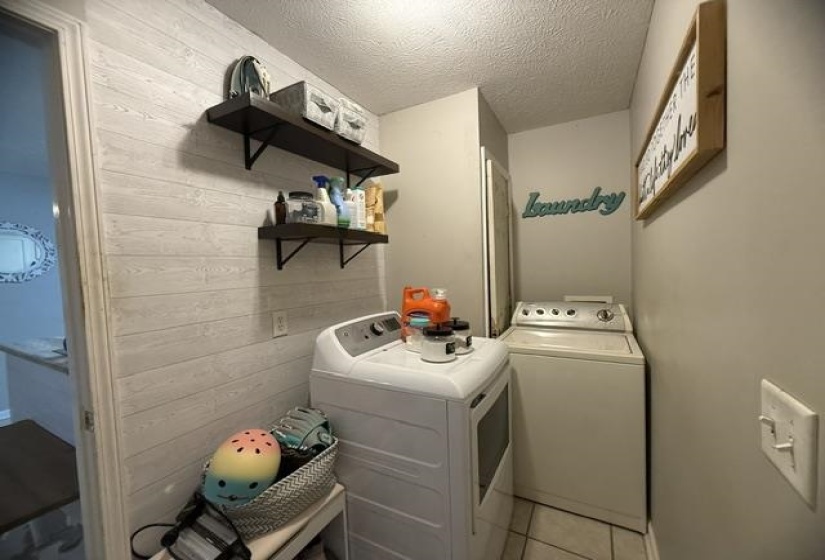 Laundry area featuring textured plank wall treatment, two floating shelves with dark wood-finish, light gray wall paint, and tiled flooring