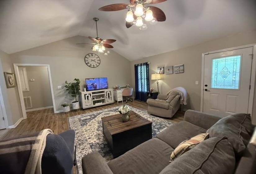 Vaulted ceiling living area featuring wood-finish flooring, two ceiling fans with integrated lighting, a white entry door with decorative glass inlay, and a doorway opening to an additional room