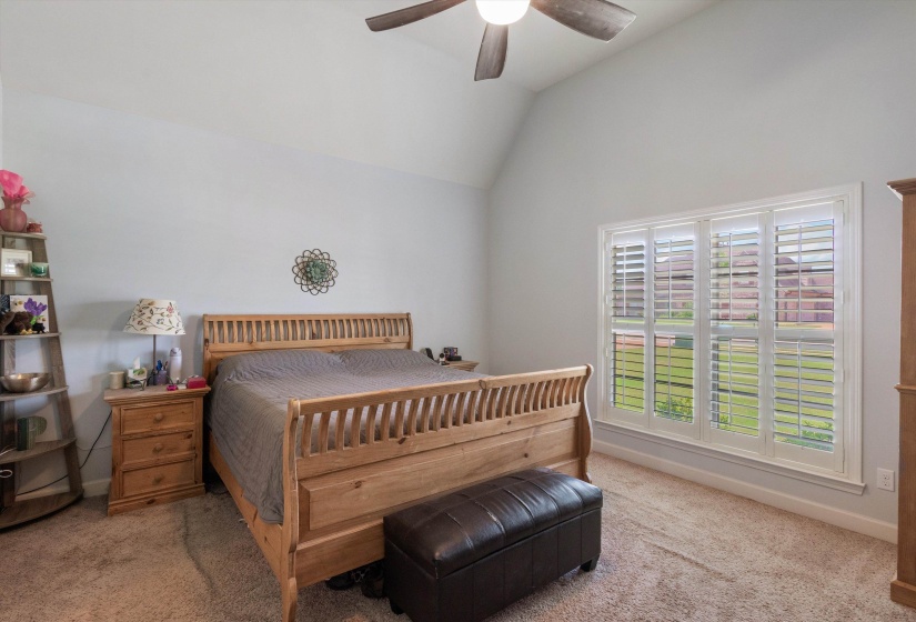 Vaulted ceiling bedroom featuring a large window with white plantation shutters, neutral carpeting, and a ceiling fan