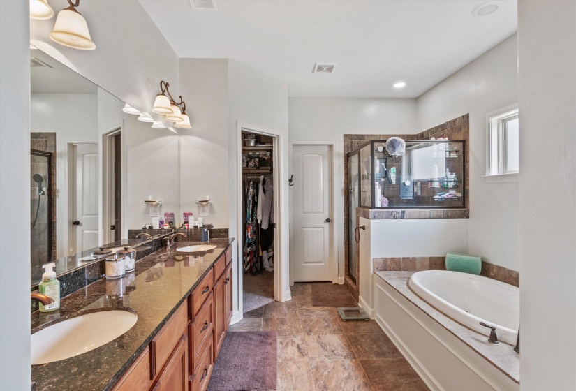 Spacious bathroom featuring a dual vanity with undermount sinks and dark countertops, a built-in soaking tub, a tiled shower enclosure with glass doors, and a separate walk-in closet
