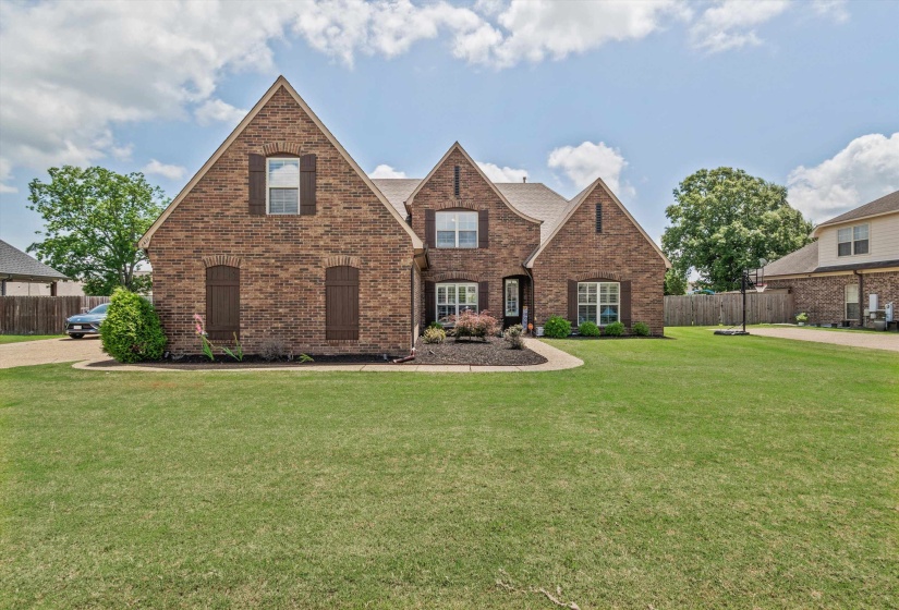 Brick exterior residence featuring multiple gables, brown window shutters, a manicured front lawn, and established landscaping