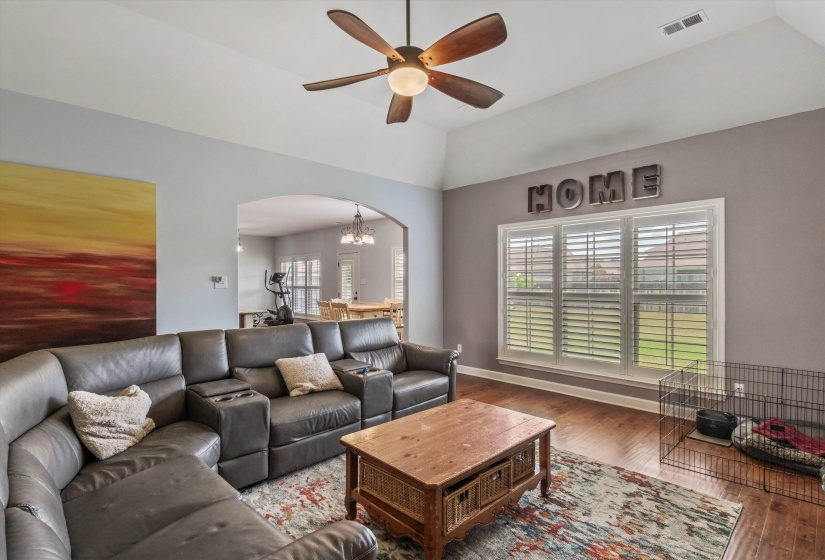 Spacious living area featuring high vaulted ceilings, wood-finish flooring, a ceiling fan with integrated lighting, a large window with white plantation shutters, and an arched entryway to an additional room