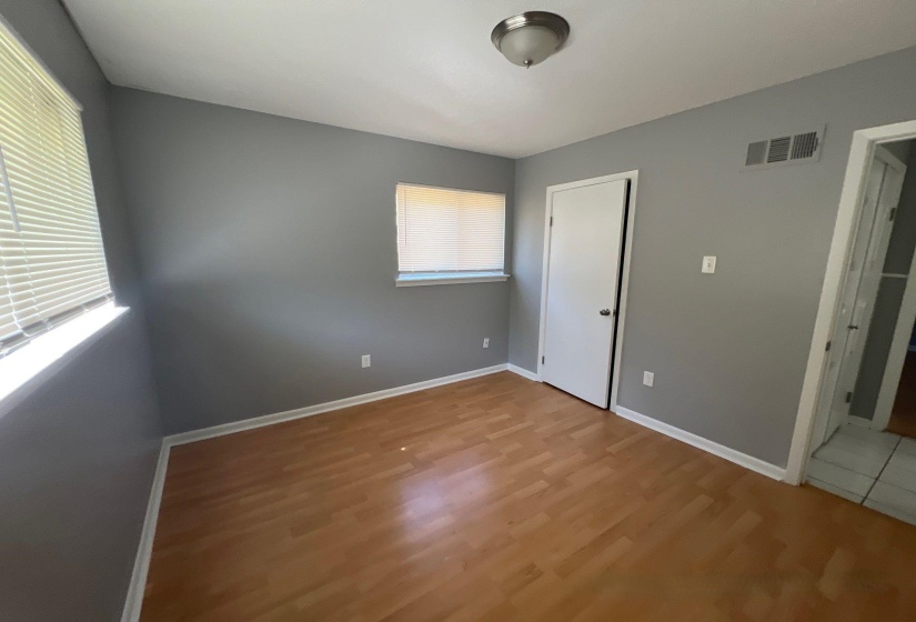 Interior room featuring wood-finish flooring, two windows with blinds, a white door with dark hardware, a ceiling-mounted light fixture, and a wall vent
