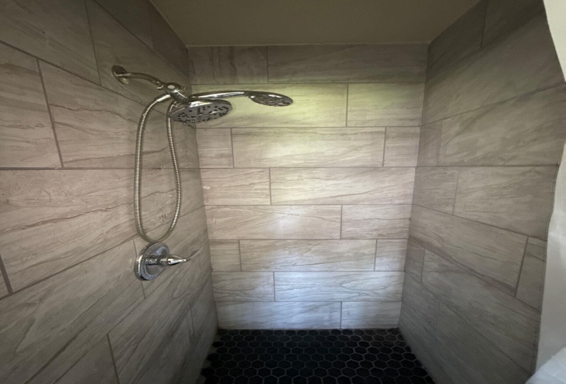 Shower stall featuring light-toned rectangular wall tiling, dark hexagonal floor tiling, and a chrome shower fixture with a handheld option