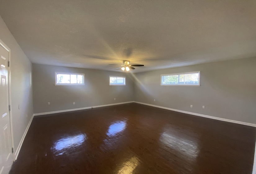 Spacious room featuring rich wood-finish flooring, light gray wall paint, white trim, multiple windows, and a ceiling fan with integrated lighting