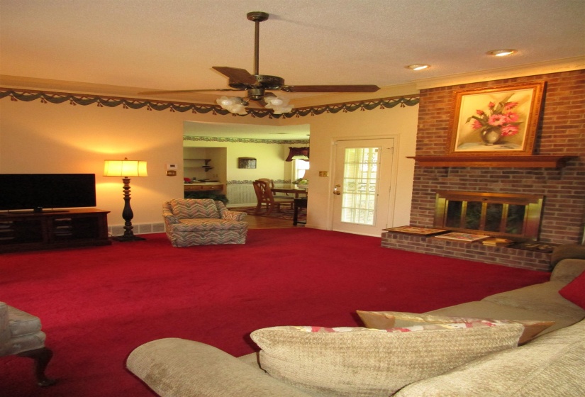 Spacious living area featuring a brick fireplace with a wood mantle, red carpeting, and a ceiling fan with integrated lighting