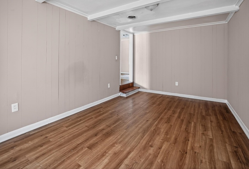 Spacious room featuring wood-finish flooring, light-toned vertical paneling, white trim, and a white painted ceiling with exposed beams