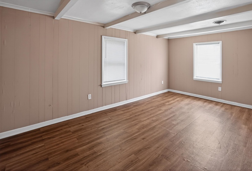 Spacious room featuring wood-finish flooring, two windows with blinds, exposed white ceiling beams, and light-colored paneled walls