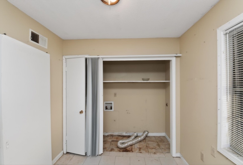 Laundry closet with tile flooring and interior shelving, featuring connections for a washing machine and dryer, along with a louvered window blind
