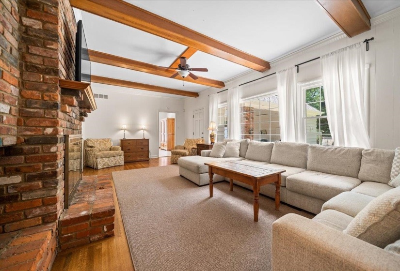 Spacious living area featuring a prominent brick fireplace, exposed ceiling beams, wet bar, multiple windows, and wood-finish flooring