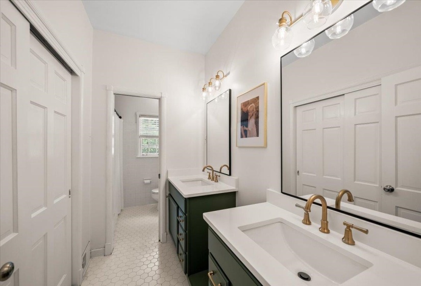 Updated bathroom featuring hexagonal floor tiling, dual vanities with deep green cabinetry, integrated sinks, and brass-finish fixtures