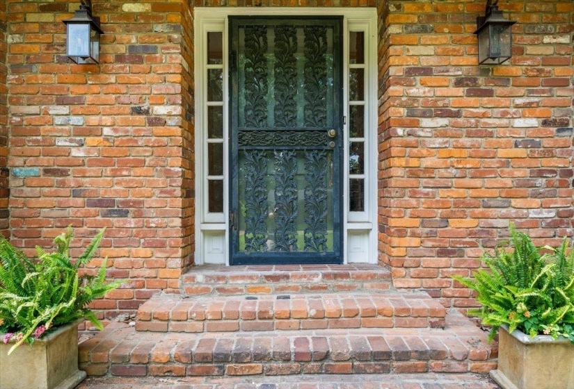 Brick facade entryway featuring an ornate black security screen door, sidelight windows, and matching exterior sconces