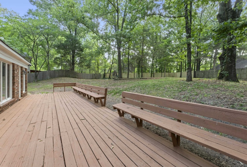 Expansive wood deck featuring built-in benches, brick exterior with multiple windows, and a generous backyard with mature trees