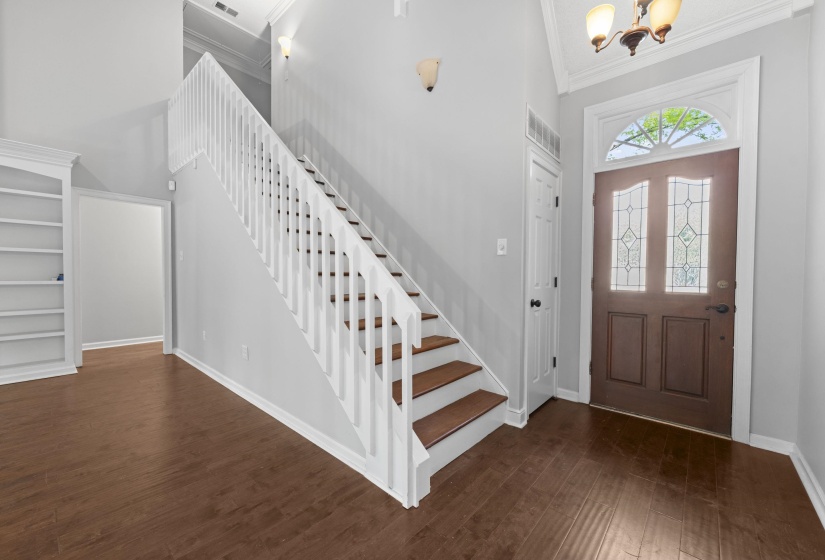 Entryway featuring wood-finish flooring, a staircase with white balusters and wood-finish treads, a built-in bookshelf, a front door with leaded glass panels and an arched transom, and a vaulted ceiling with crown molding