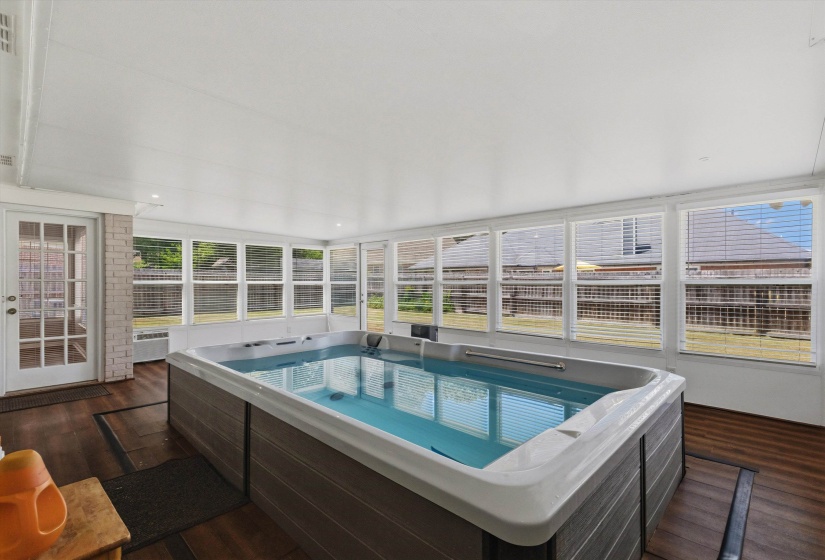Indoor swim spa area featuring wood-finish flooring, white brick accent wall, white-framed windows with blinds, and a white paneled ceiling with recessed lighting