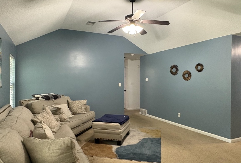 Spacious living area featuring a vaulted ceiling with a ceiling fan, light blue wall paint, and beige carpeting