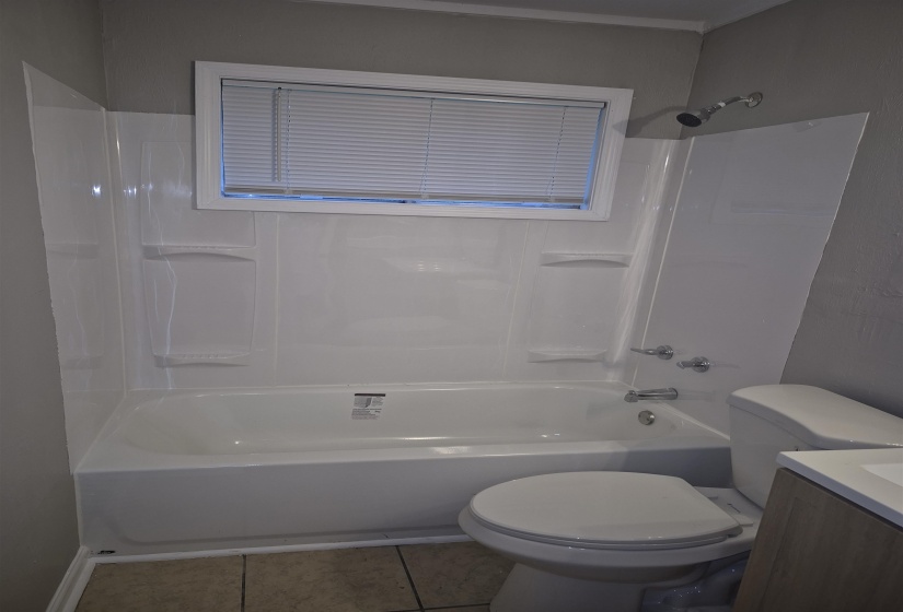 Bathroom featuring a white bathtub with integrated shower, white shower surround, chrome shower head, and a window with blinds