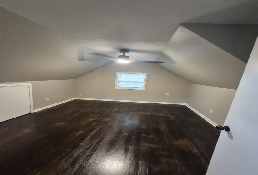 Attic room featuring dark wood-finish flooring, a window with blinds, and a built-in ceiling fan with integrated lighting