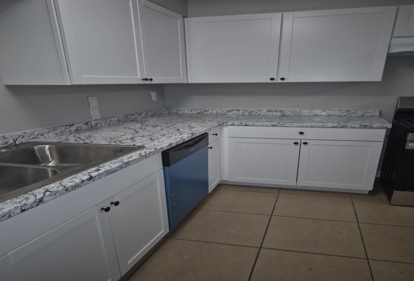 Kitchen featuring white shaker-style cabinetry with black hardware, mottled light-toned countertops, a double-basin stainless steel sink, and a built-in dishwasher