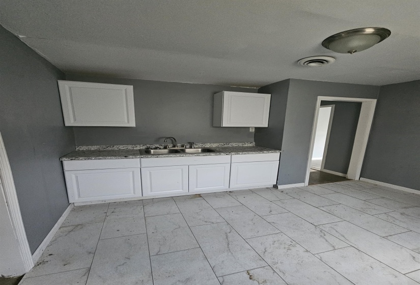 Kitchenette featuring light gray tile flooring, white shaker-style cabinetry, and a double basin stainless steel sink with a speckled countertop
