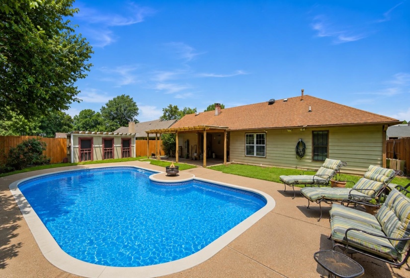 Freeform swimming pool with a surrounding concrete patio, covered patio area, and a private wooden fence. Some features are AI-enhanced.