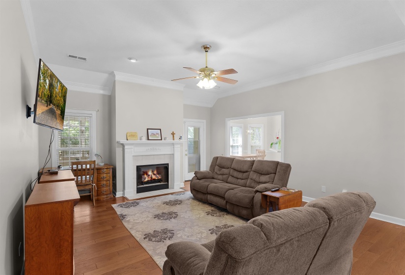 Living room featuring wood-finish flooring, a white mantel fireplace, a ceiling fan, crown molding, and a visible doorway to an additional room