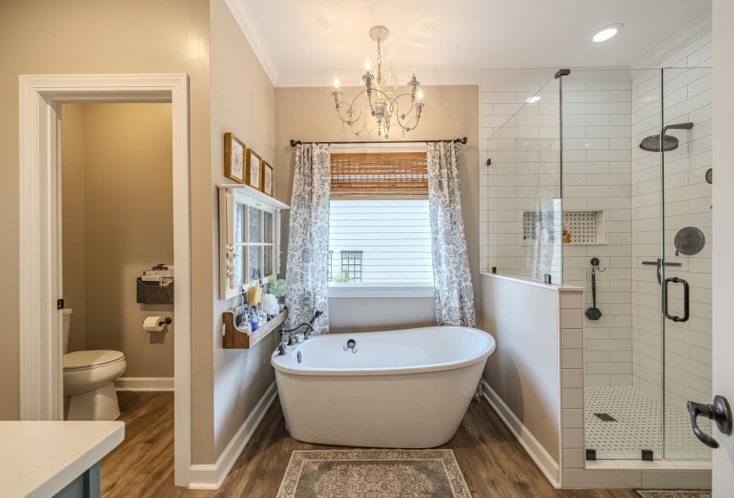Freestanding soaking tub with a chandelier overhead, glass-enclosed shower with subway tile surround, wood-finish flooring, and a separate water closet