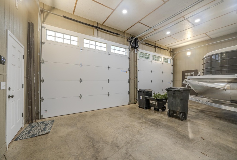 Spacious garage featuring two white overhead doors with transom windows, exposed ceiling beams, recessed lighting, and a durable concrete floor