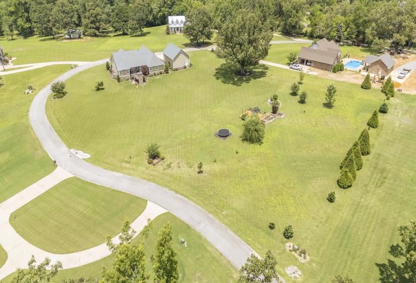 Expansive green lawn surrounds multiple properties, featuring a winding paved driveway, mature trees, and a distant swimming pool