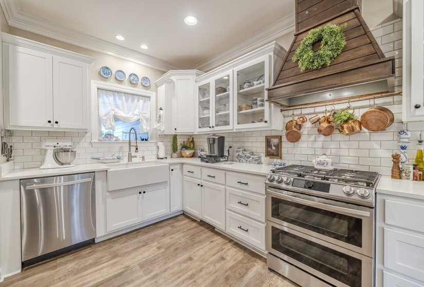 Bright kitchen featuring white shaker cabinetry, stone countertops, a farm sink, stainless steel appliances, and wood-finish flooring