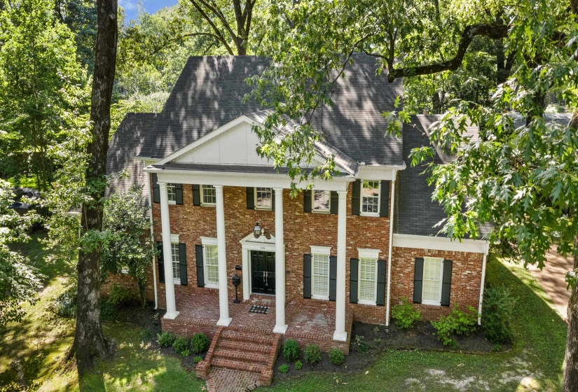 Brick two-story colonial exterior featuring a portico with white columns, a dark shingled roof, and dark shutters