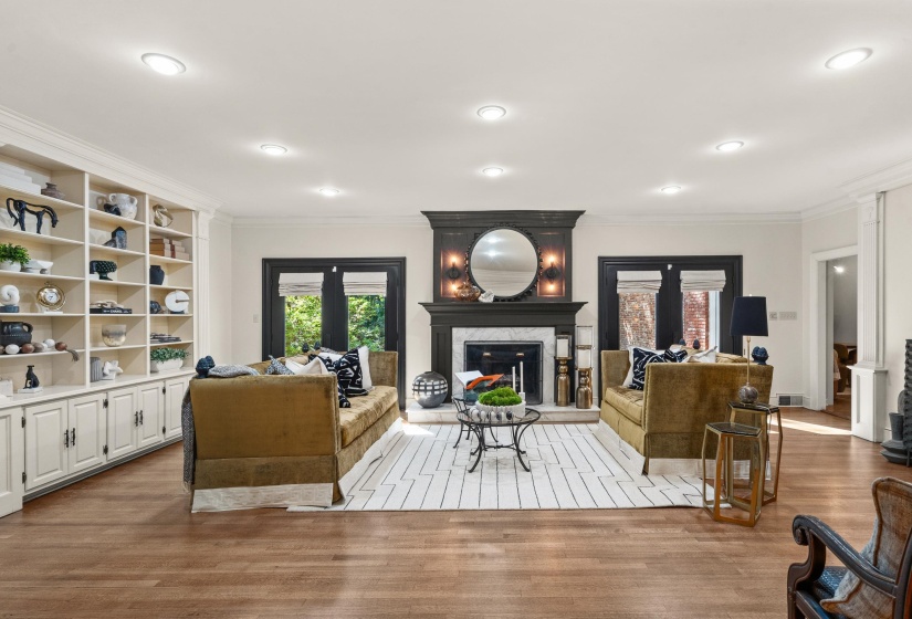 Spacious living area featuring rich wood-finish flooring, a prominent fireplace with a dark wood mantel and marble surround, built-in shelving and lower cabinetry, recessed lighting, and double French doors