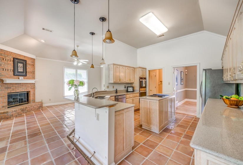 Open-concept kitchen and living area featuring vaulted ceilings, terracotta tile flooring, a brick fireplace, pendant lighting, and light wood cabinetry with light-colored countertops