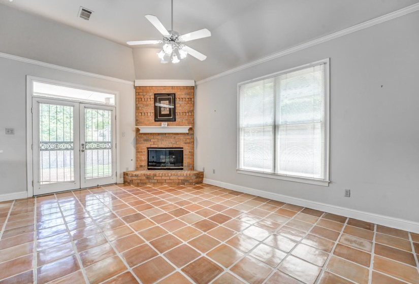Spacious room featuring terracotta-style tile flooring, a corner brick fireplace with a white mantel, and a ceiling fan with light fixture