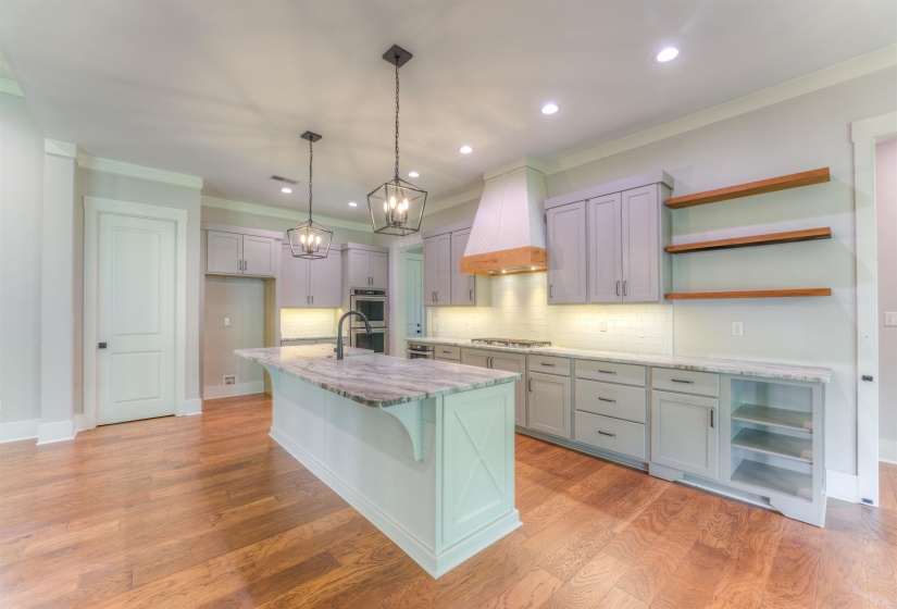 Spacious kitchen featuring an island with a stone countertop, light gray cabinetry, built-in double ovens, a wood-accented range hood, and open wood shelving