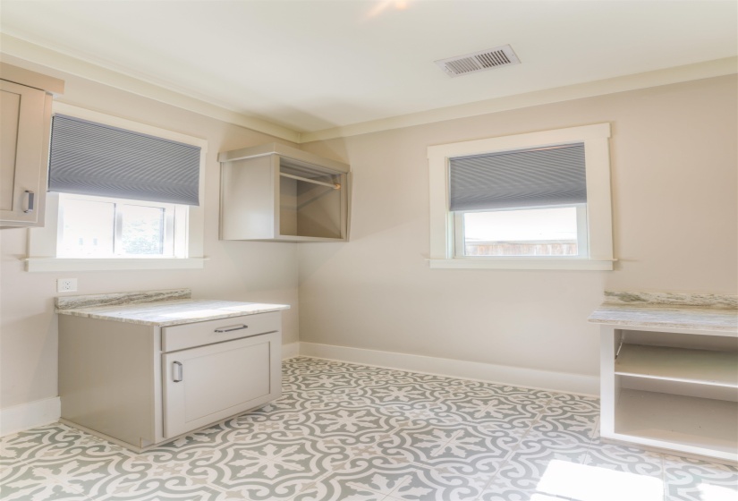Laundry room featuring patterned floor tiles, light gray cabinetry, stone-finish countertops, two windows with blinds, and crown molding
